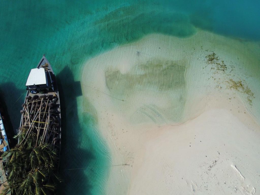 Aerial view of a boat loaded with mature palms beside a white sandbank in a turquoise Maldivian lagoon